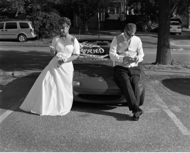 Black and White Image of a Bride and Groom sitting on the hood of a car looking at imaginary phones