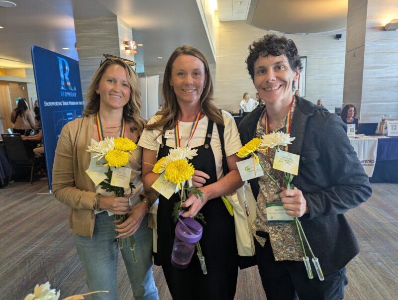 Three conference attendees stand together smiling in a hotel conference lobby, each holding yellow and white flowers with handwritten appreciation notes attached. They are wearing colorful ribbon lanyards and conference name badges, celebrating connection and recognition during a community-focused event. Booths and attendees are visible in the background.
